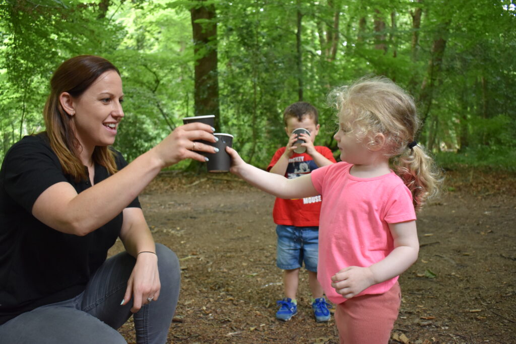 What We Do: Wellies in the woods photo of a woman holding cups with two young children in outdoor setting.