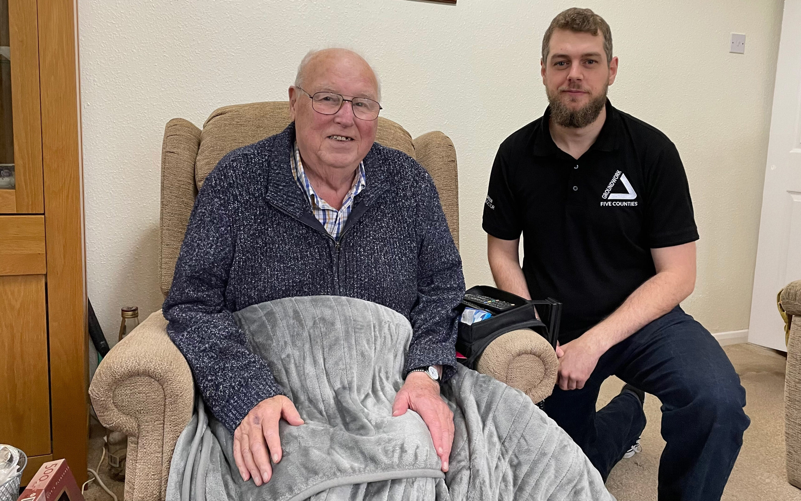 Elderly man in chair with grey blanket and man kneeling next to him in black shirt with white Groundwork logo.