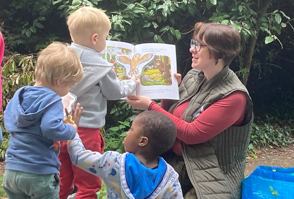 What We Do: Woman reading from picture book with children around, in outdoor setting.